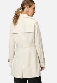 Beige Trenchcoat mit tailliertem Schnitt, Gürtel in der Taille, Schulterklappen und geknöpften Manschetten, aus glattem Stoff gefertigt.