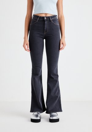 Person trägt hoch taillierte, dunkel ausgestellte Jeans und silberne Plateauschuhe, steht vor einem schlichten hellen Hintergrund.