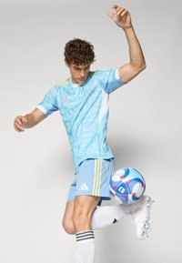 Camiseta de fútbol azul claro con patrones abstractos, detalles en blanco y tres rayas amarillas. Pantalones cortos azul claro y botas blancas, pateando un balón multicolor.