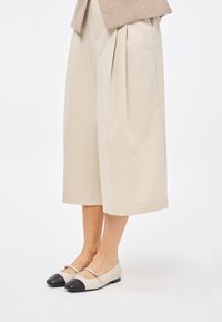 Scarpe basse beige con punta nera, caratterizzate da una sottile fascia sul dorso del piede e una superficie in pelle liscia.