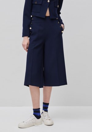 Nogi w granatowych, szerokich, skróconych spodniach z złotymi guzikami, w paskowane granatowo-niebieskie skarpetki oraz białe trampki na jednolitym tle.