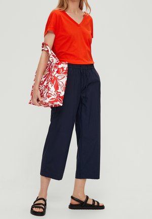 Frau trägt leuchtend orangefarbenes V-Ausschnitt-Shirt, marineblaue weite, kurze Hosen und schwarze Sandalen, hält eine rote und weiße Blumentasche.