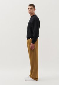 Sweatshirt nero a maniche lunghe con polsini a costine, abbinato a pantaloni giallo senape e sneakers bianche. La silhouette è rilassata e moderna.