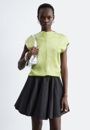 Femme portant un haut vert clair à manches courtes et une jupe noire évasée, tenant un petit sac à main métallique argenté sur l'épaule, fond neutre.