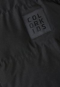 Tela negra con una textura acolchada y un parche de goma que dice "COLOR KIDS" en letras bold, centrado en la tela.