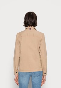 Béžová džínová bunda s límcem, detailem na zadní části a manžetami na knoflíky. Obsahuje pruhovaný textilní akcent na výstřihu.