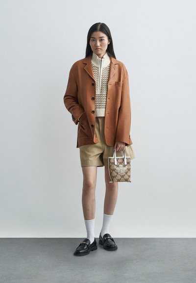 Kobieta w brązowej kurtce, beżowym swetrze z dzianiny, khaki szortach, czarnych mokasynach, trzymająca małą torebkę w wzory, stojąca na szarym podłożu przy białej ścianie.
