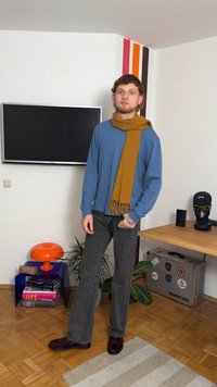 Cămașă albastră cu mâneci lungi, eșarfă muștar cu franjuri, blugi evazați gri, pantofi închiși la culoare. Stă într-un interior modern cu plante și decorațiuni.