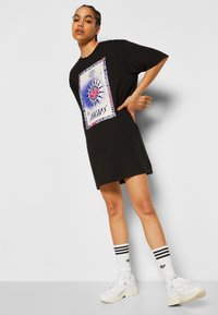 Black oversized t-shirt dress with a colorful graphic print, short sleeves, and loose fit. Paired with white sneakers and striped socks.