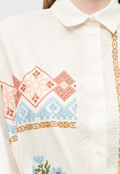 Λευκό πουκάμισο με κουμπιά και παραδοσιακά κεντημένα γεωμετρικά μοτίβα σε πορτοκαλί, μπλε και καφέ κατά μήκος του ώμου και του στήθους.