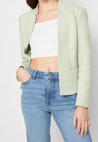 Blazer vert clair texturé à motif chevrons, ouvert sur le devant et coupe ajustée, associé à un jean bleu taille haute et un crop top blanc.