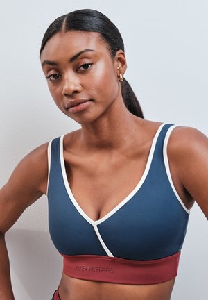 Femme aux cheveux foncés attachés, portant un brassière de sport bleue et rouge avec un liseré blanc sur un fond uni.