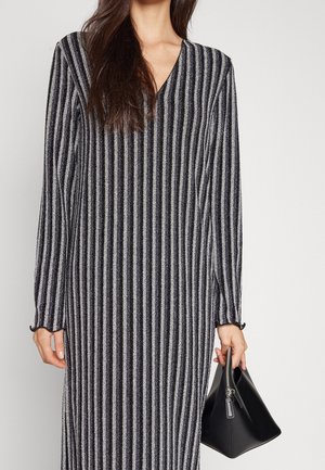 Kobieta w długiej sukience w pionowe paski w kolorze czarno-szarym, trzymająca w prawej ręce czarną trójkątną torebkę na suwak.