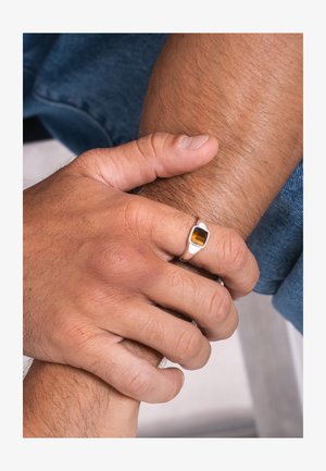 Zwei Hände, die ein Handgelenk umfassen, eine Hand trägt einen silbernen Ring mit einem rechteckigen Bernstein, im Hintergrund ist Jeansstoff zu sehen.