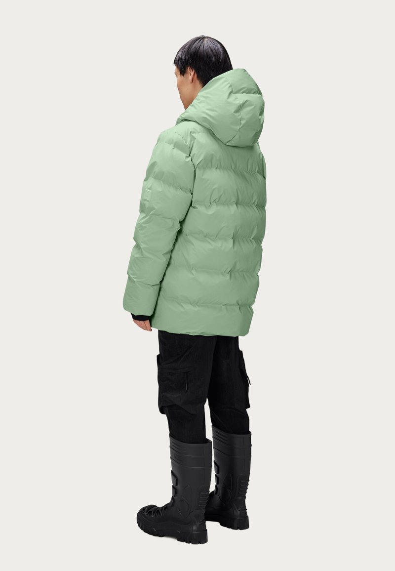 Veste verte en doudoune avec une capuche, présentant une texture lisse et des segments cousus ; associée à un pantalon cargo noir et des bottes noires jusqu'aux genoux.