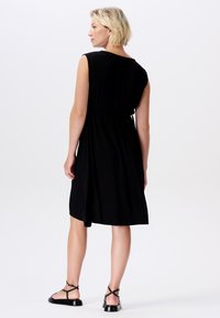 Schwarzes knielanges Kleid mit lockerer Passform, ausgestattet mit kurzen Ärmeln und einem Kordelzug in der Taille, aus strukturiertem Stoff gefertigt. Tragen mit flachen Sandalen.