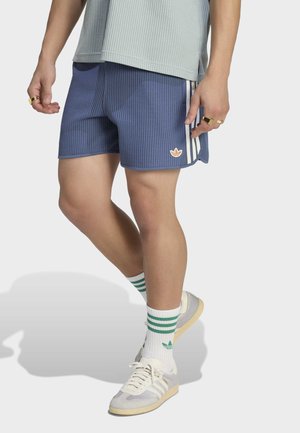 Persona con pantalones cortos azules texturizados con rayas blancas y logo de Adidas, zapatillas blancas, calcetines blancos con rayas verdes, de pie.