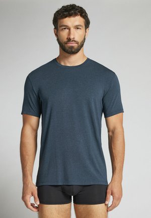 Mann mit lockigem Haar und Bart, der ein schlichtes dunkelblaues kurzärmeliges T-Shirt und schwarze Boxershorts trägt, steht vor einem neutralen Hintergrund.