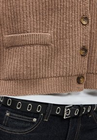 Κοντινό πλάνο καφέ πλεκτής ζακέτας με κουμπιά πάνω από λευκό πουκάμισο και μαύρη ζώνη με ασημί οπές σε σκουρόχρωμο τζιν.