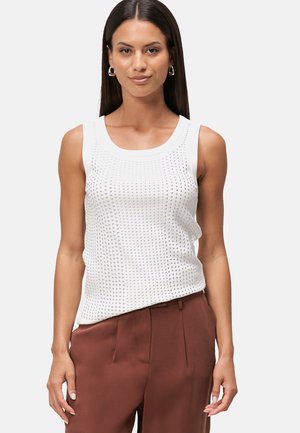 Top sans manches blanc avec un motif de tricot texturé, doté d'un col rond et de bords lisses, associé à un pantalon marron ajusté.