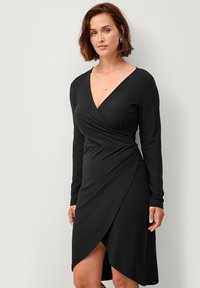 Zwarte wikkeljurk met lange mouwen heeft een diepe V-halslijn, een cinched taille en een diagonale zoom die een elegante silhouet creëert.