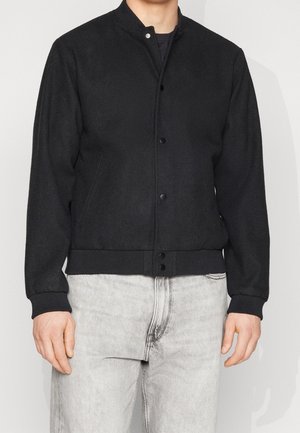 Homme portant une veste bomber noire boutonnée avec poignets et ourlet côtelés, associée à un jean en denim gris clair.