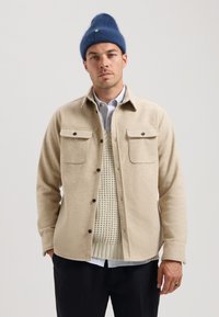 Beige flannel skjortejakke med to brystlommer, sorte knapper, båret over en tekstureret cremesweater med en blus med blå striber under.