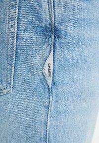 Svijetloplave traperice s blagom izblijedjelom teksturom. Imaju malu tkanu oznaku s natpisom "CHASIN'" blizu šava džepa.