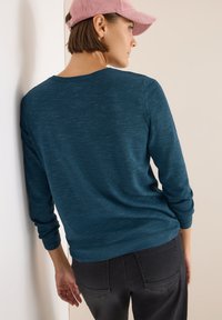 Maglione in maglia teal con maniche lunghe, scollo rotondo e vestibilità morbida. Indossato con pantaloni neri e un berretto di velluto a coste rosa.