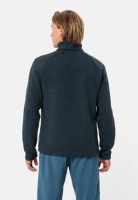 Jachetă din fleece albastru închis, cu un căptușit înalt, textură încrețită și mâneci lungi. Prezintă un material contrastant la umeri și o etichetă verde la margine.