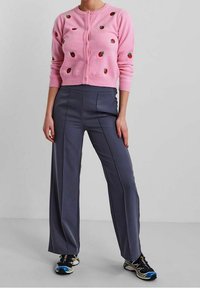 Person som bär en rosa cardigan med broderade jordgubbar, mörka högmidjade vida byxor och svarta sportskor med färgglada detaljer.