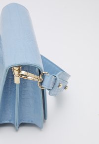 Błękitna torebka wykonana z błyszczącego, teksturowanego materiału przypominającego skórę krokodyla. Posiada złote zapięcie i regulowany pasek.