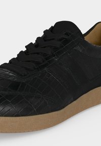 Baskets basses noires avec tige en cuir texturé, lacets noirs et semelle en caoutchouc marron clair sur fond blanc.