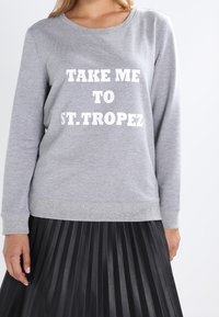 Gray sweatshirt with long sleeves and crew neck featuring white text "TAKE ME TO ST. TROPEZ." Worn over a black pleated skirt.