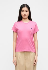 Γυναίκα με μακριά σκούρα μαλλιά φοράει ένα ροζ κοντομάνικο μπλουζάκι με ένα μικρό μπλε λογότυπο στο στήθος και μπεζ παντελόνες με φαρδιά πόδια.