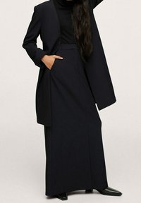 Zwarte op maat gemaakte kostuum bestaande uit een lange blazer met brede revers en een bijpassende hoge taillebroek. Glad textuur, minimale details en een ontspannen pasvorm.