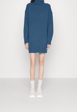 Femme portant une robe pull à col roulé bleue côtelée associée à des bottines blanches, debout contre un fond clair uni.