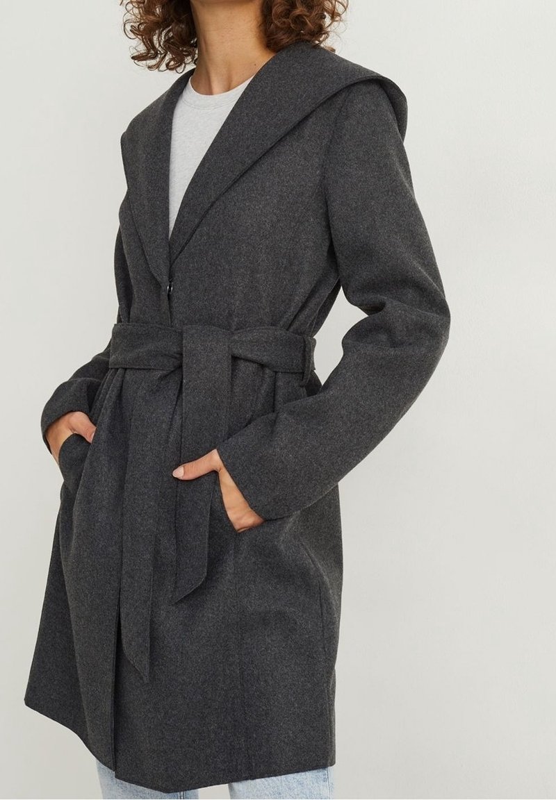 Femme portant un manteau en laine gris foncé ceinturé sur une chemise gris clair, les mains dans les poches, sur un fond clair uni.