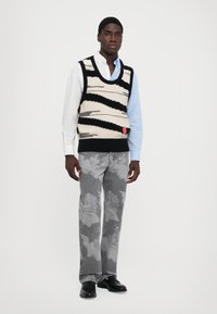 Débardeur en tricot avec un design en zigzag noir et crème, superposé sur une chemise bleu clair. Pantalon gris avec un motif d'ange fané. Chaussures noires.
