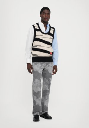 Gebreide tanktop met zwart en romige zigzagpatroon, gelaagd over een lichtblauw shirt. Grijze broek met een vervaagd engelpatroon. Zwarte schoenen.