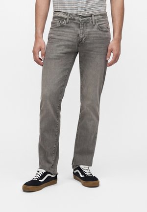 Homme portant un jean gris délavé, une chemise à carreaux rentrée et des baskets noires avec des lacets blancs et des semelles en gomme, debout devant un fond uni.