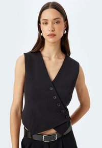 Haut sans manches noir enroulé avec décolleté en V, présentant cinq boutons noirs sur le côté et un tissu lisse et structuré. Accompagné d'une ceinture.
