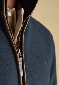 Nahaufnahme einer marineblauen Fleecejacke mit braun lederbesetztem Reißverschluss, der teilweise geöffnet ist und ein beigefarbenes Hemd mit Kragen darunter zeigt.