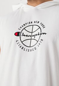 Bílá bezrukávová mikina s kapucí s předním potiskem, na kterém se nachází kulaté logo basketbalu a text "Champion New York Established 1919" v černé a červené.