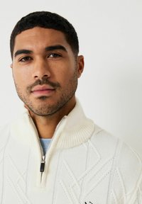 Adīta pus-izmēra džemperis krēmkrāsā ar ribotu apkakli un teksturētu dimanta rakstu. Iekļauts metāla rāvējslēdzis ar melnu vilkšanas cilpu.