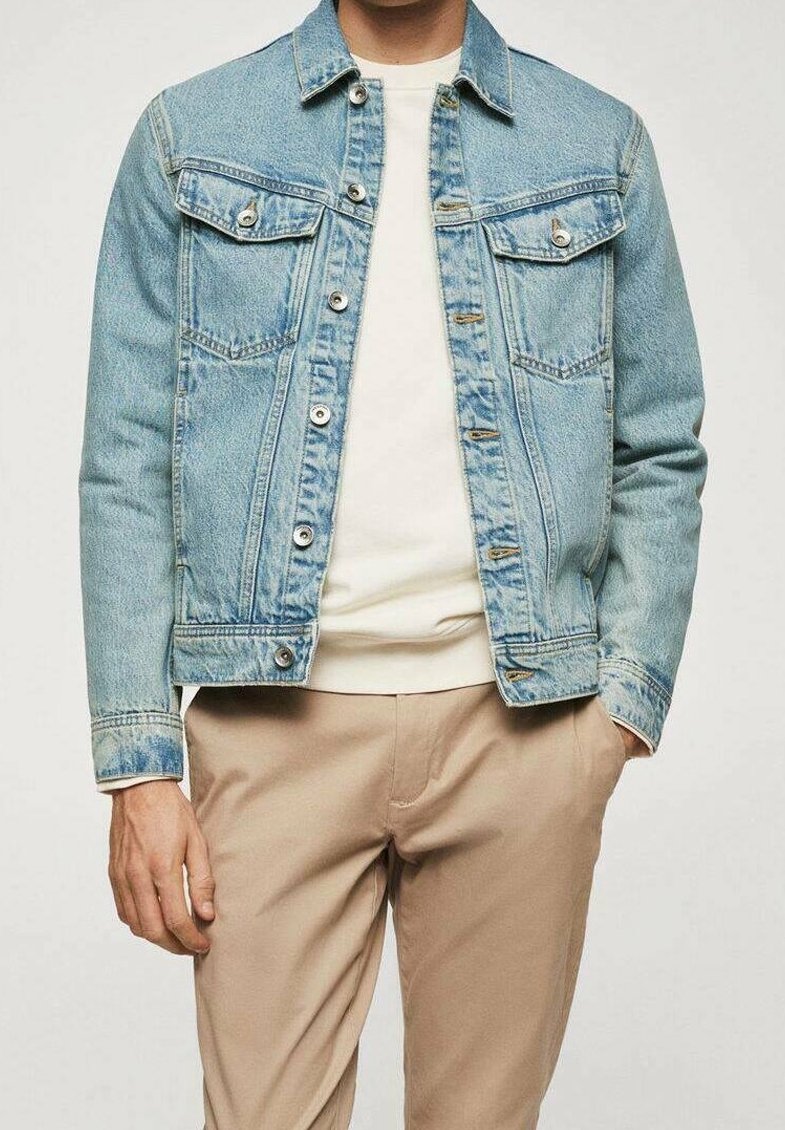 Homme portant une veste en jean bleu clair sur une chemise blanche et un pantalon beige, une main dans la poche, sur un fond uni.