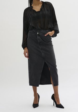Femme portant un chemisier noir transparent, une jupe midi en denim gris foncé taille haute avec une fente avant et des escarpins noirs à bouts pointus.
