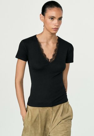 Femme aux cheveux foncés en chignon lisse porte un haut noir à col en V avec bordure en dentelle et un pantalon plissé beige, debout les mains dans les poches.