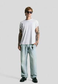 Witte oversized t-shirt met opgerolde mouwen, lichtblauwe baggy jeans, zwarte zonnebril en zwarte instappers met een sleutelhanger-detail.