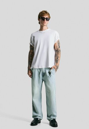 Weißes übergroßes T-Shirt mit aufgerollten Ärmeln, hellblaue baggy Jeans, schwarze Sonnenbrille und schwarze Slipper mit Schlüsselanhänger-Detail.